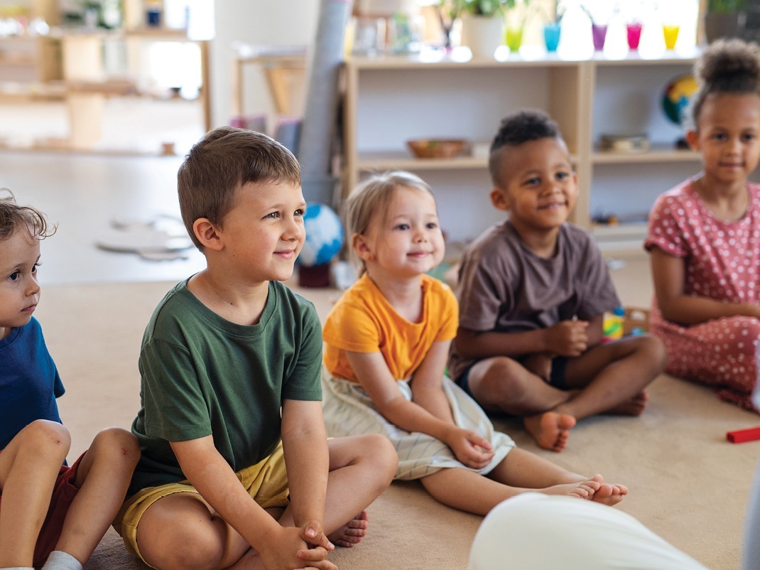 Cinq jeunes enfants sont assis sur un tapis moelleux et sourient à leur éducatrice.
